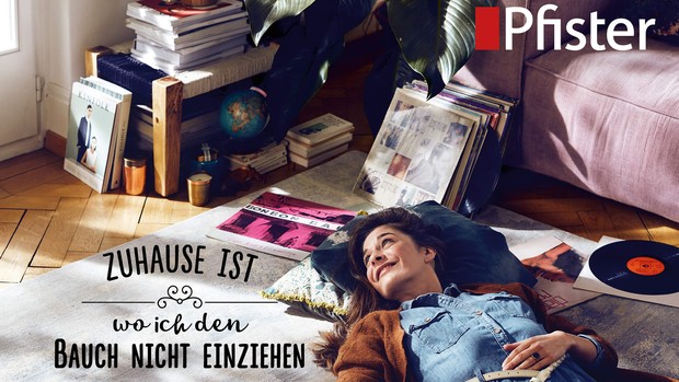 Glückliche Frau liegt auf Boden vor dem Sofa mit Schriftzug "Zuhause ist, wo ich den Bauch nicht einziehen muss"